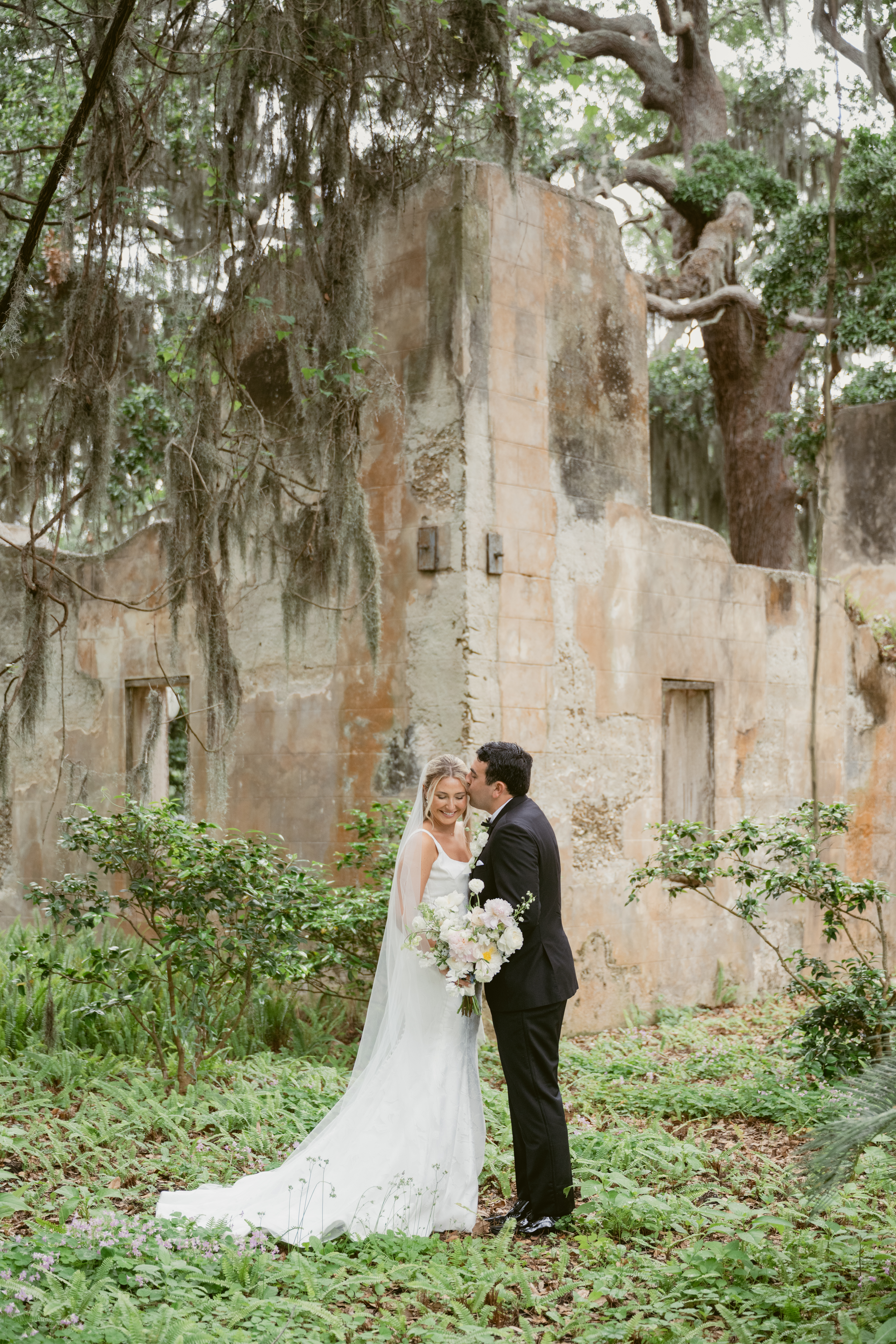 Bride and groom stand in front of romantic stone ruins surrounded by greenery; the groom kisses the bride on the temple as she smiles, holding a bouquet of soft white and blush flowers.