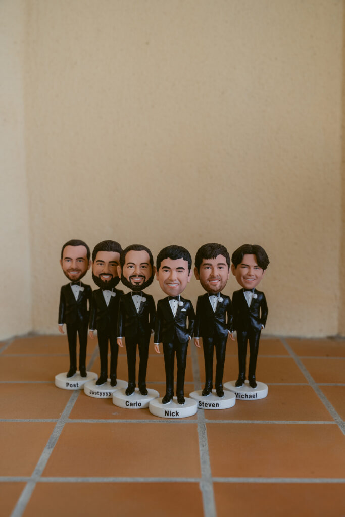 Custom groomsmen bobblehead figurines lined up on a tile floor, each labeled with names like Nick, Steven, Carlo, and Michael.