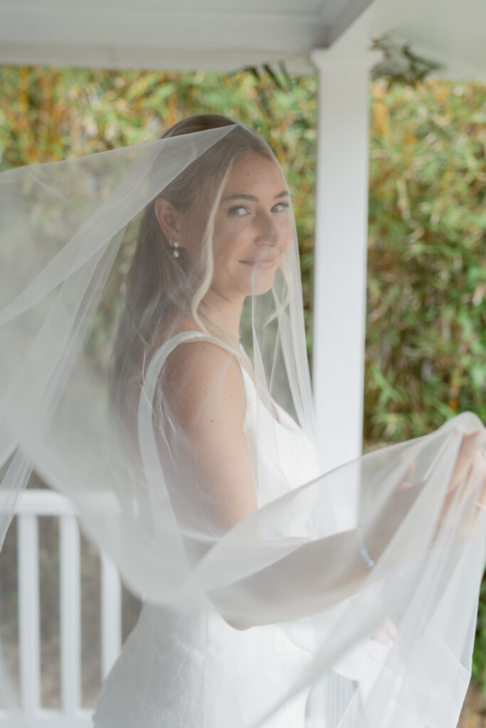 Bride standing on a porch with soft light filtering through her veil, smiling sweetly just before her Georgia wedding ceremony.