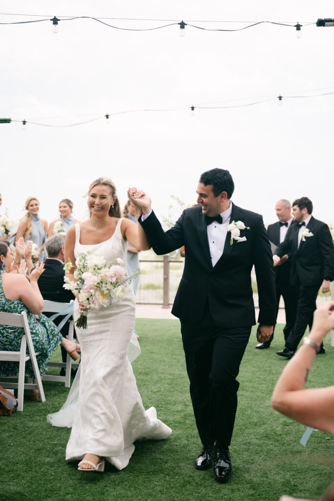 Bride and groom walk down the aisle smiling as guests cheer during outdoor ceremony.