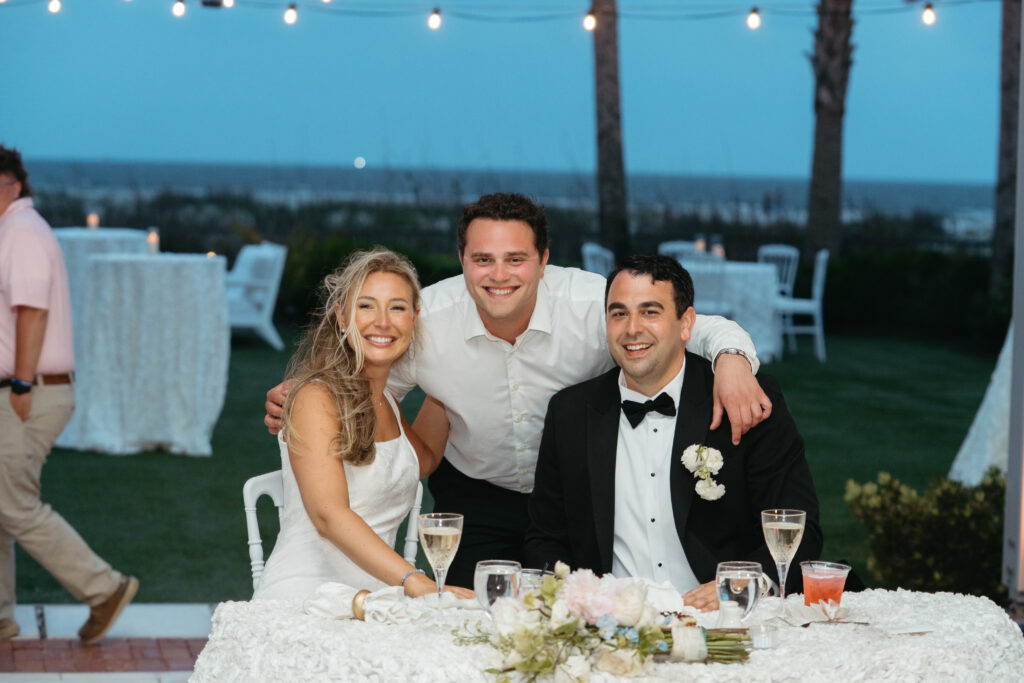 Bride and groom sit at their sweetheart table smiling with a guest, during the outdoor evening reception at The King and Prince Resort.