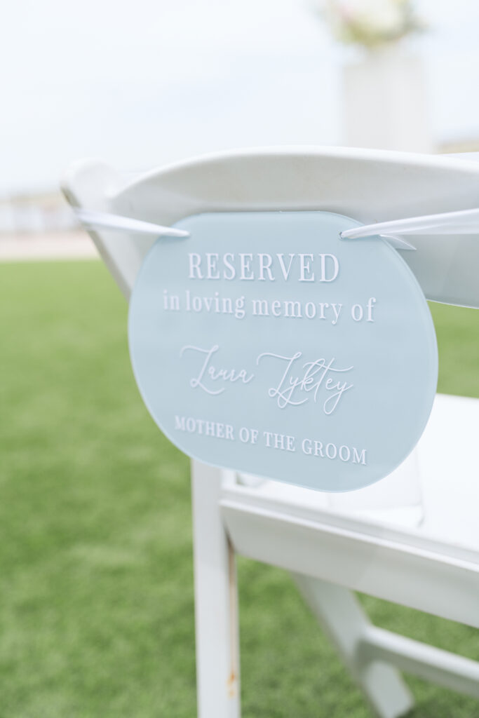 A white ceremony chair with a soft blue sign that reads, "RESERVED in loving memory of Laura Lyktey, MOTHER OF THE GROOM," honoring a loved one at a beachside wedding.