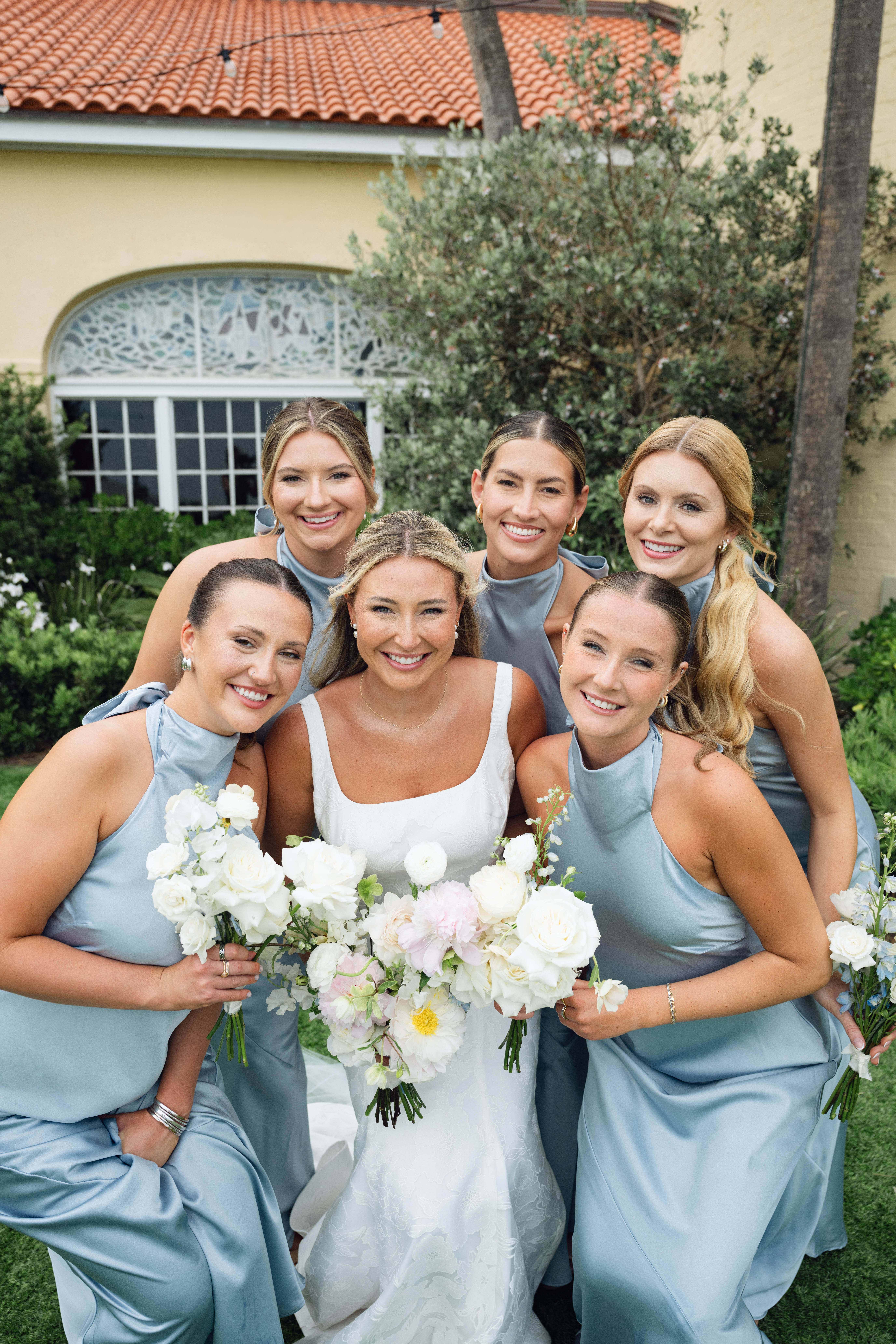 Bride and bridesmaids in light blue dresses holding white and blush bouquets, smiling outdoors.
