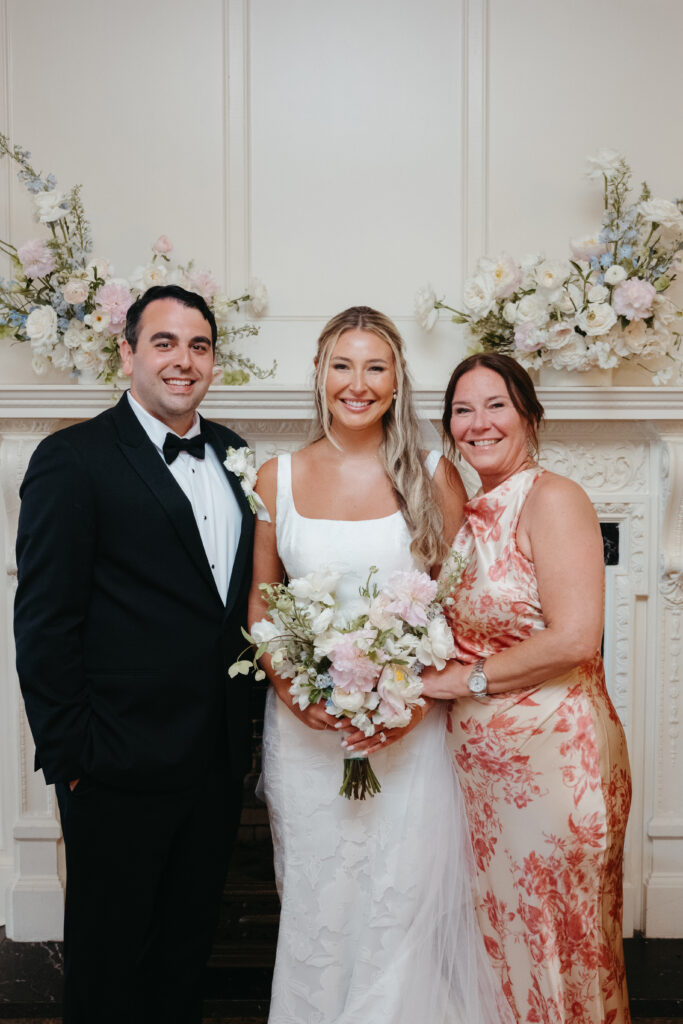 Bride and groom smiling with a guest in front of a floral-decorated fireplace indoors.