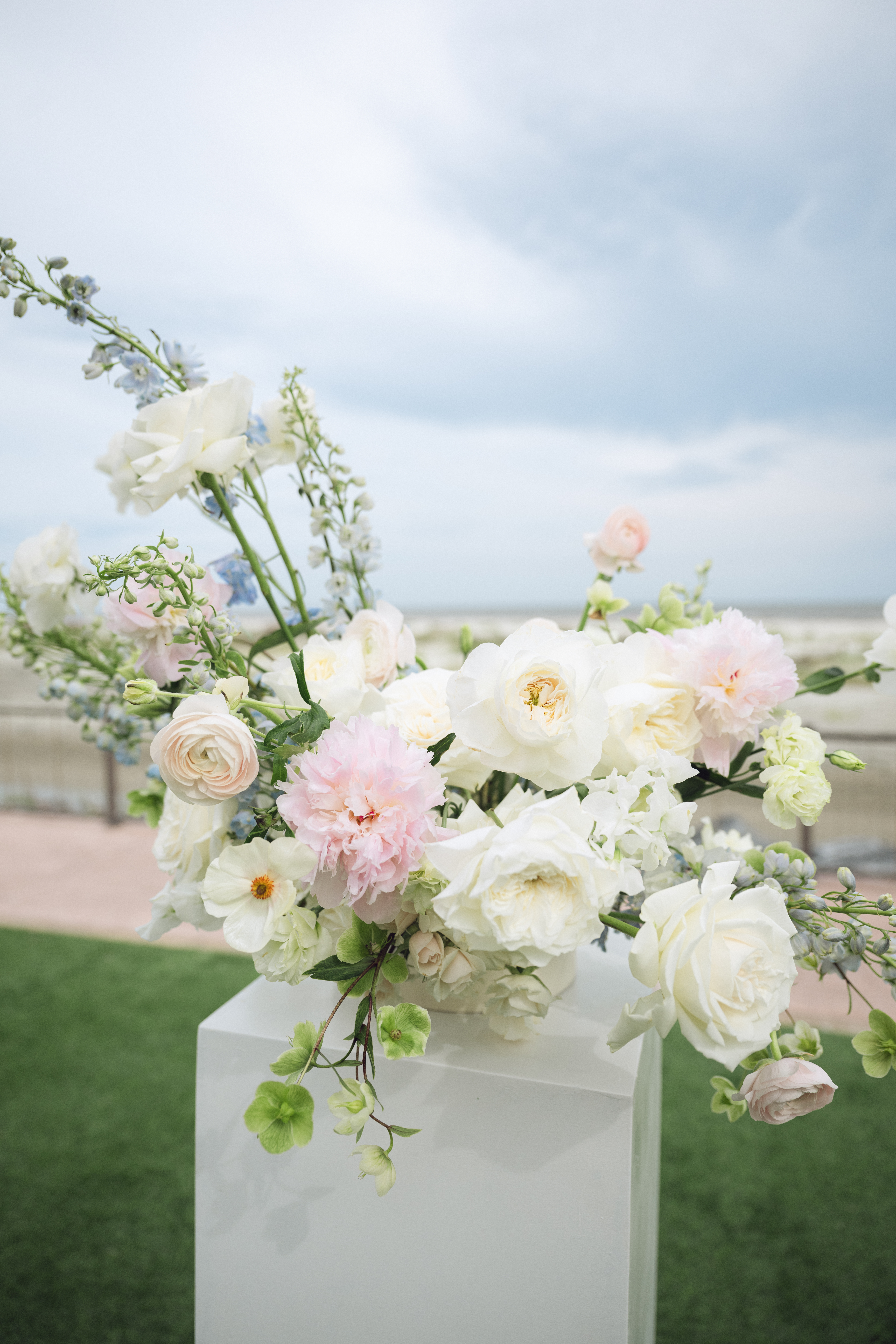 A romantic beachside floral arrangement featuring soft pastel blooms including white roses, blush peonies, and delicate greenery, displayed on a modern white pedestal.