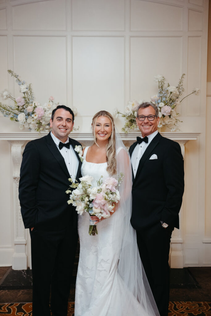 Bride and groom smiling with bride's father in front of elegant floral fireplace indoors.