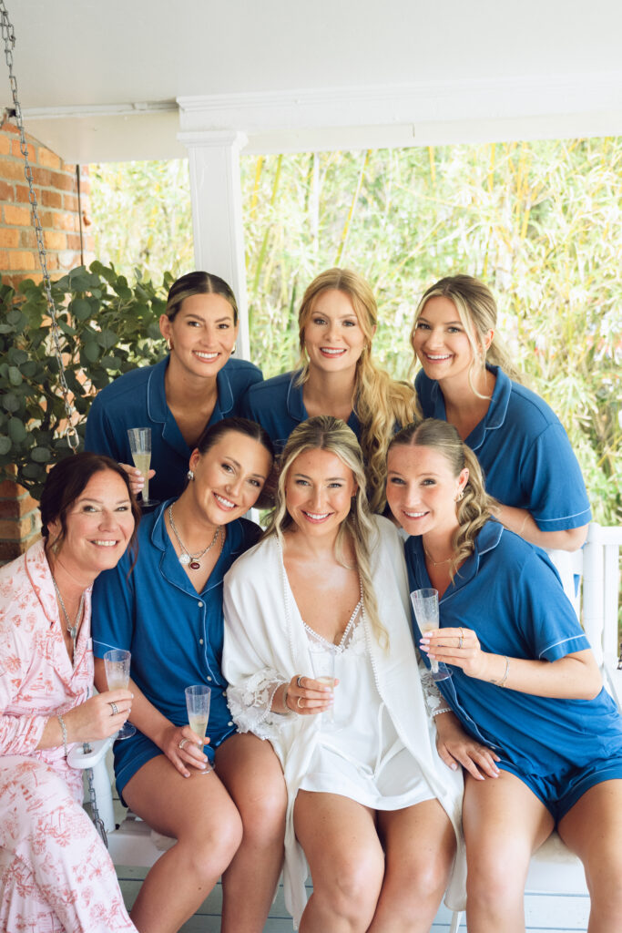 Bride in white robe surrounded by bridesmaids in matching blue pajamas, smiling and holding champagne glasses on the porch during a Georgia wedding morning.