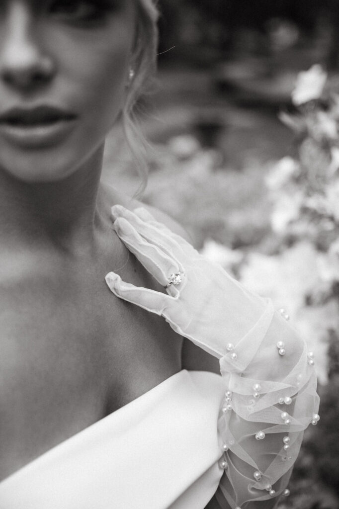 Black and white close-up of a bride wearing a pearl-detailed glove and off-the-shoulder gown, gently resting her hand near her collarbone to show her ring.