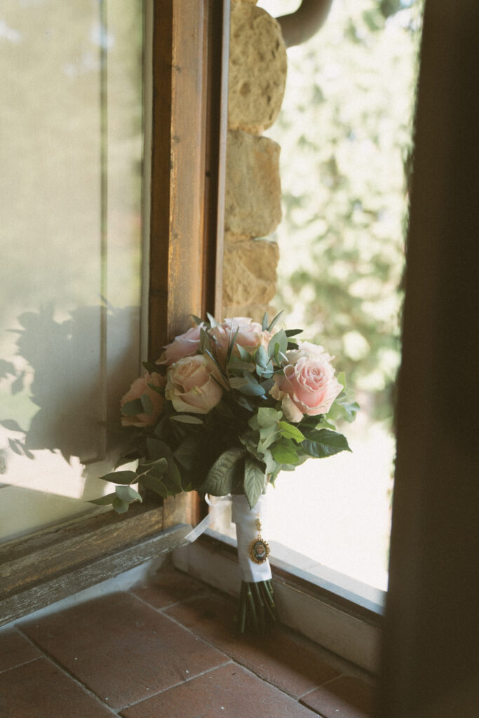 Bridal bouquet of soft pink roses and greenery resting against a sunlit window, wrapped with white ribbon and a vintage gold charm.