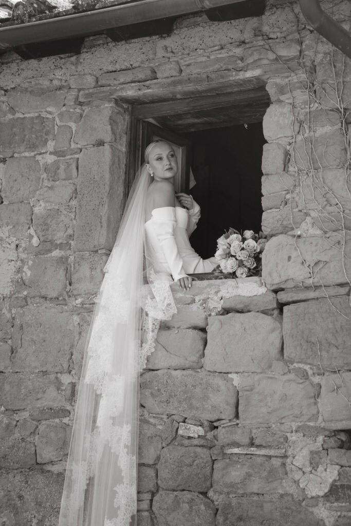 Black and white photo of a bride in an off-the-shoulder gown and long lace veil, posing thoughtfully in a rustic stone window with a bouquet nearby.