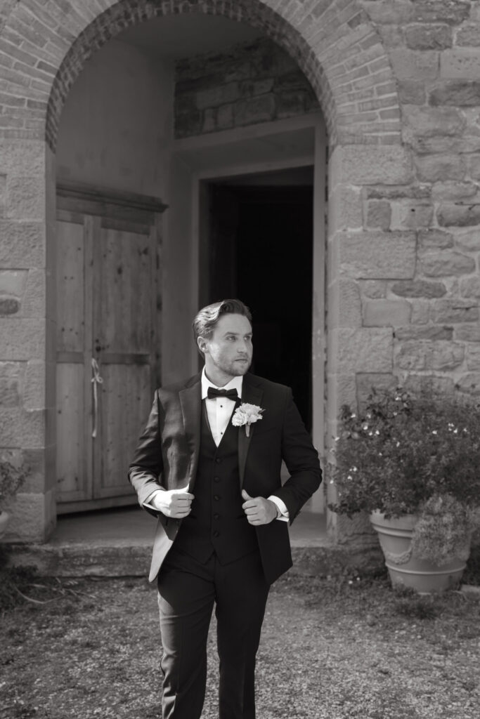 Black and white photo of a groom adjusting his suit jacket while confidently walking outside a stone building with an arched doorway.