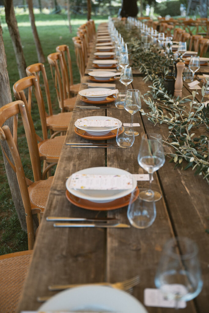 Long rustic wooden table set for an outdoor wedding reception, with neatly arranged place settings, glassware, and a garland of greenery down the center.