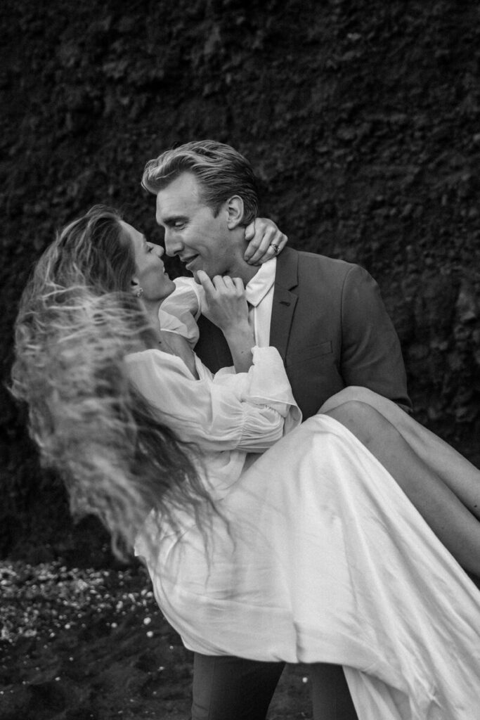 Black and white photo of a groom holding his bride in his arms on a beach, both laughing and gazing at each other as her hair blows in the wind.