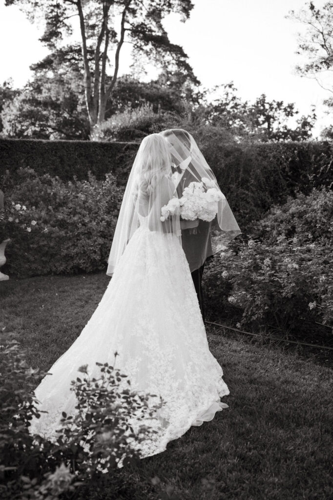 Black and white photo of a bride and groom sharing a quiet moment in a garden, standing face to face under her veil as she holds a bouquet.