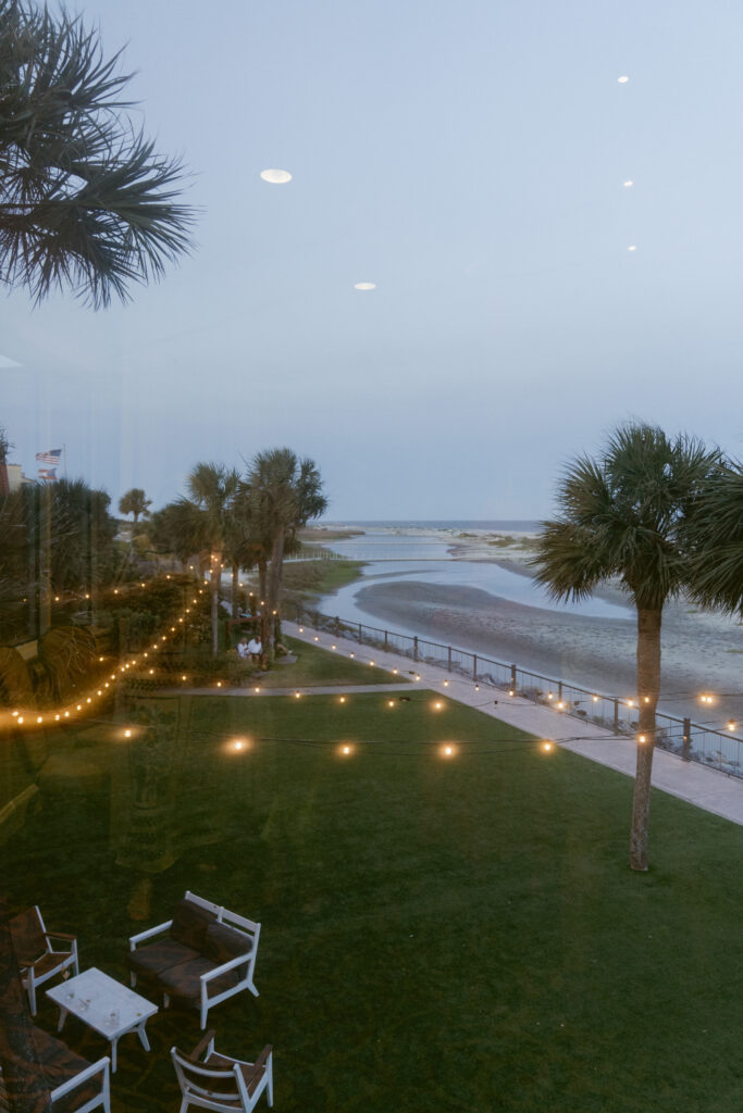 View of a coastal lawn at The King and Prince Resort with string lights, palm trees, and ocean in the distance during twilight.