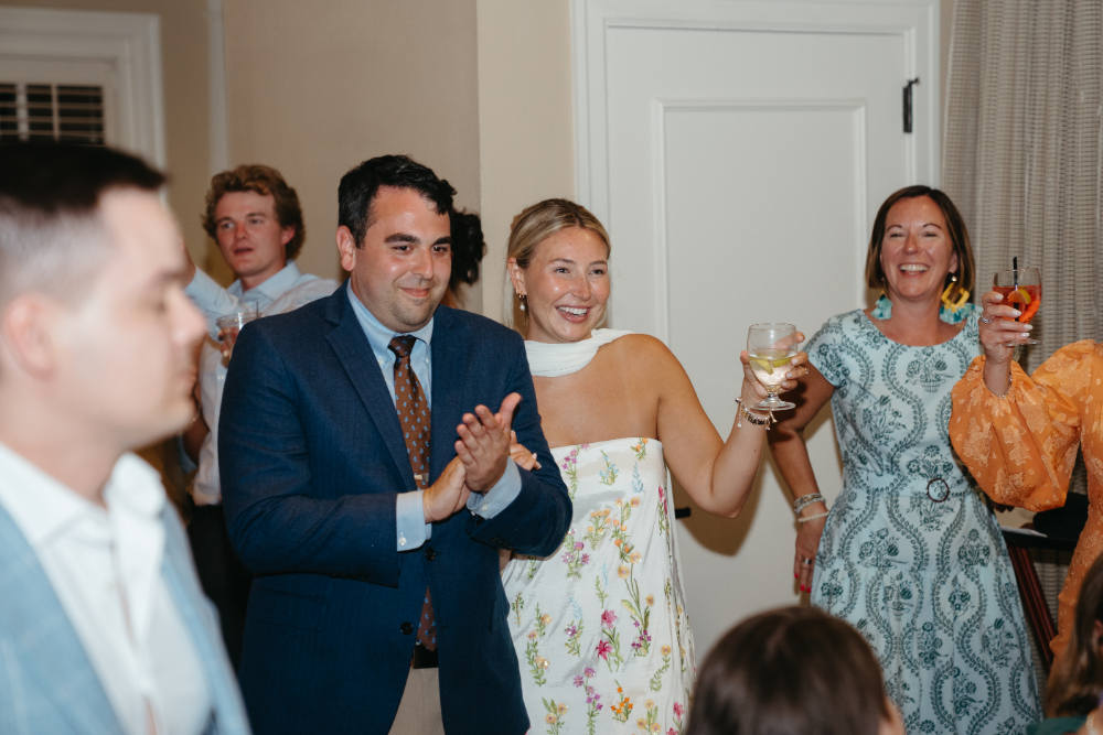 Bride in a floral dress and groom in a navy suit smiling and raising glasses during the rehearsal dinner toast.