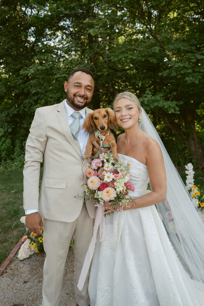 Bride and groom smile with their dog and bouquet, standing together in a sunny garden setting.