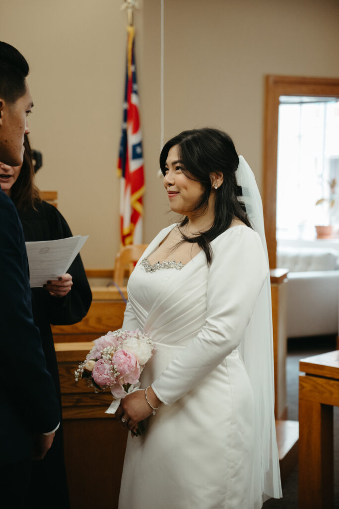 Bride smiles at groom during their intimate wedding ceremony inside a courthouse with natural light.