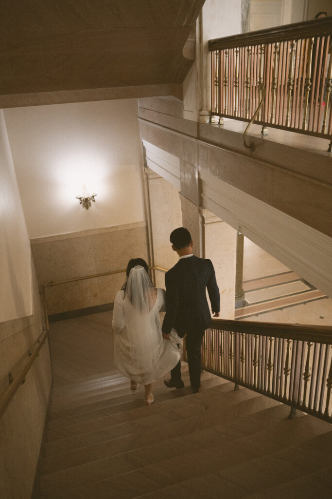 Bride and groom walk down a grand staircase inside a historic courthouse, hand in hand.