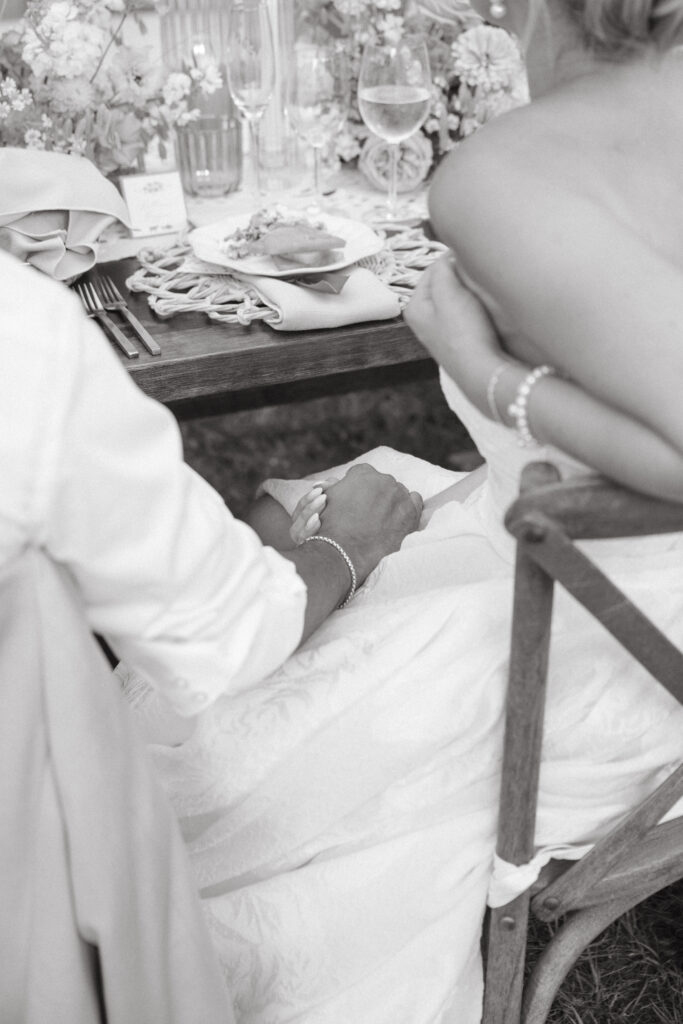 black and white photo of a bride and groom holding hands while listening to the speeches at their wedding