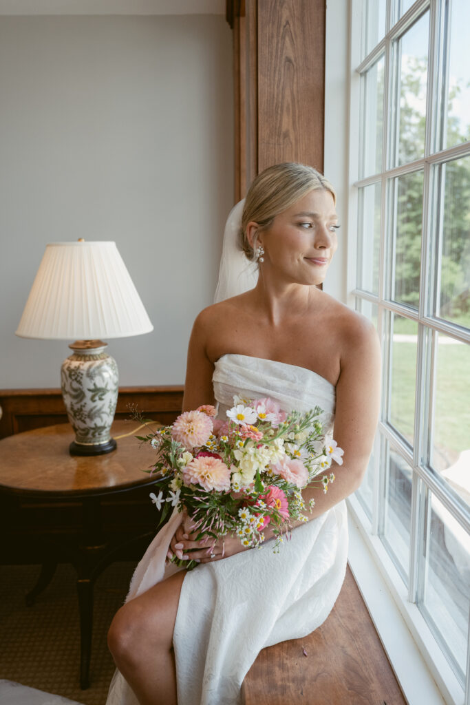 Bride sits by window holding bouquet, captured in soft light by Cincinnati wedding photographer.