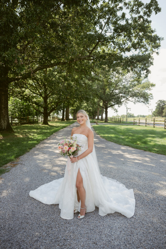 Bride stands smiling on a tree-lined path holding bouquet-Wedding at Meshewa House