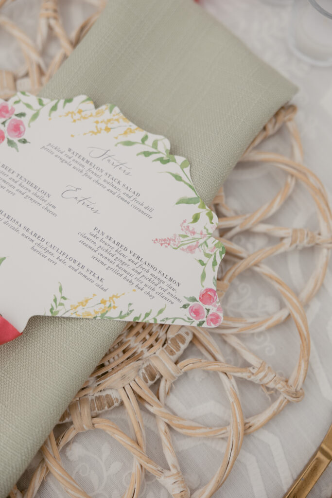 Close-up of a wedding menu card with floral illustrations, placed on a green napkin and woven charger at a beautifully styled wedding reception table.