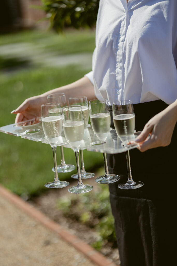 Server holding a clear tray of champagne glasses during outdoor wedding cocktail hour.