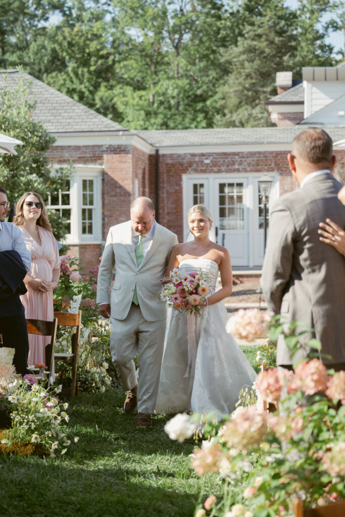 Bride walks down the flower-lined aisle with her father during a sunny outdoor ceremony.