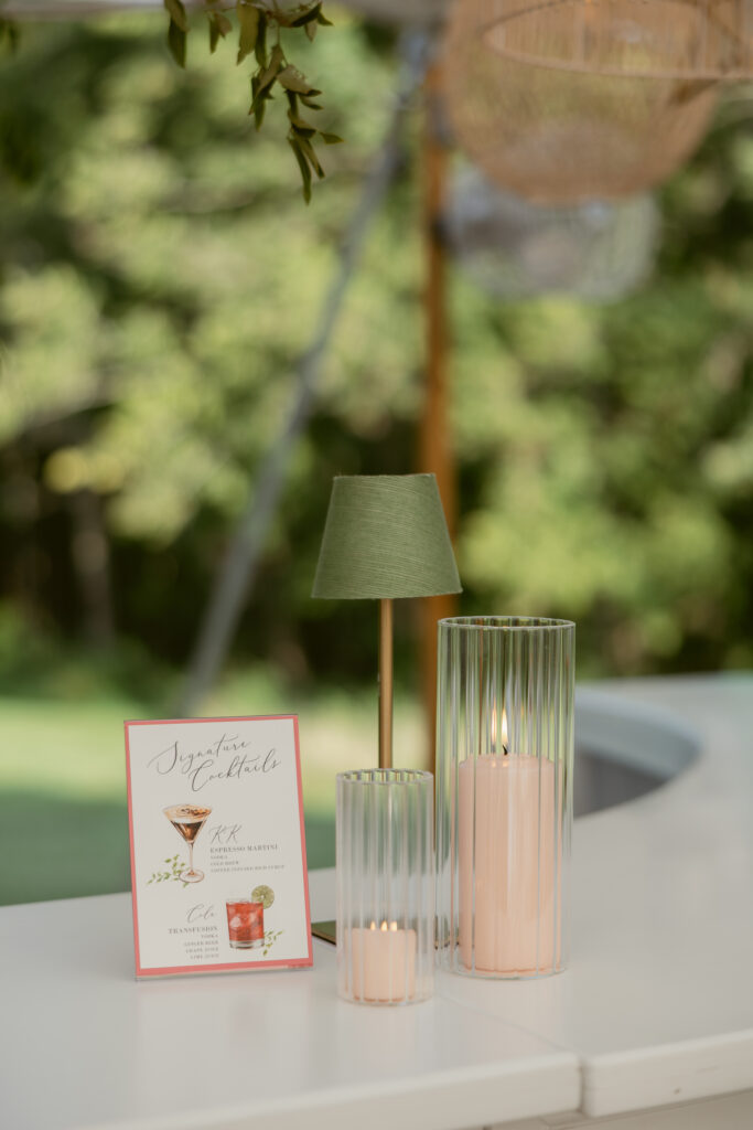 Close-up of signature cocktail sign, candles, and green mini lamp on modern white wedding bar.