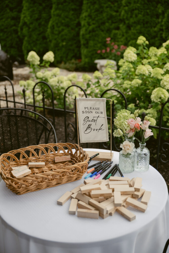 Outdoor guest book table featuring wooden Jenga blocks, colorful pens, and simple floral arrangements.