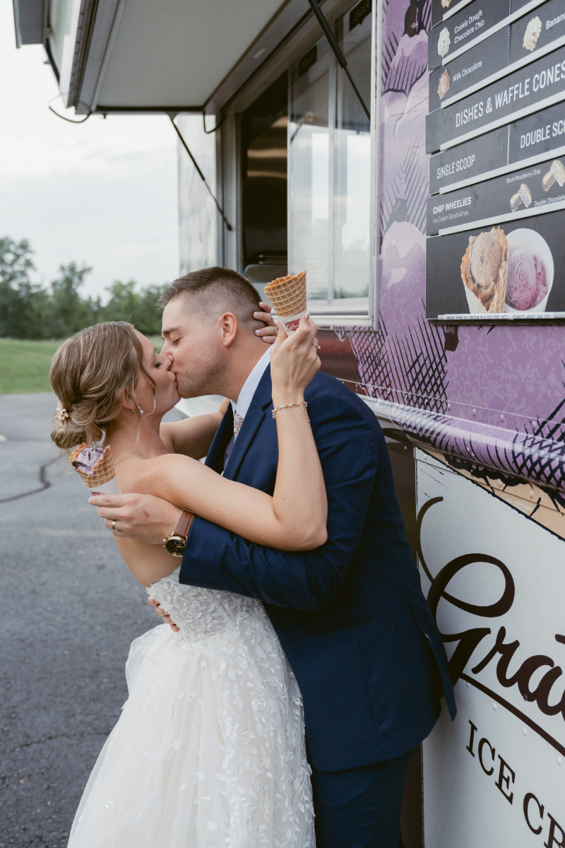 Bride and groom share a kiss while holding ice cream cones in front of a colorful food truck.