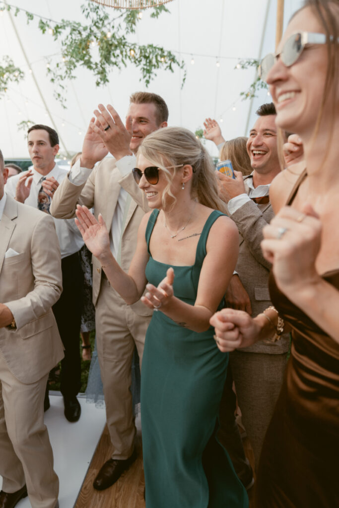 Wedding guests in formal attire laughing and dancing together under a tented reception with greenery above.