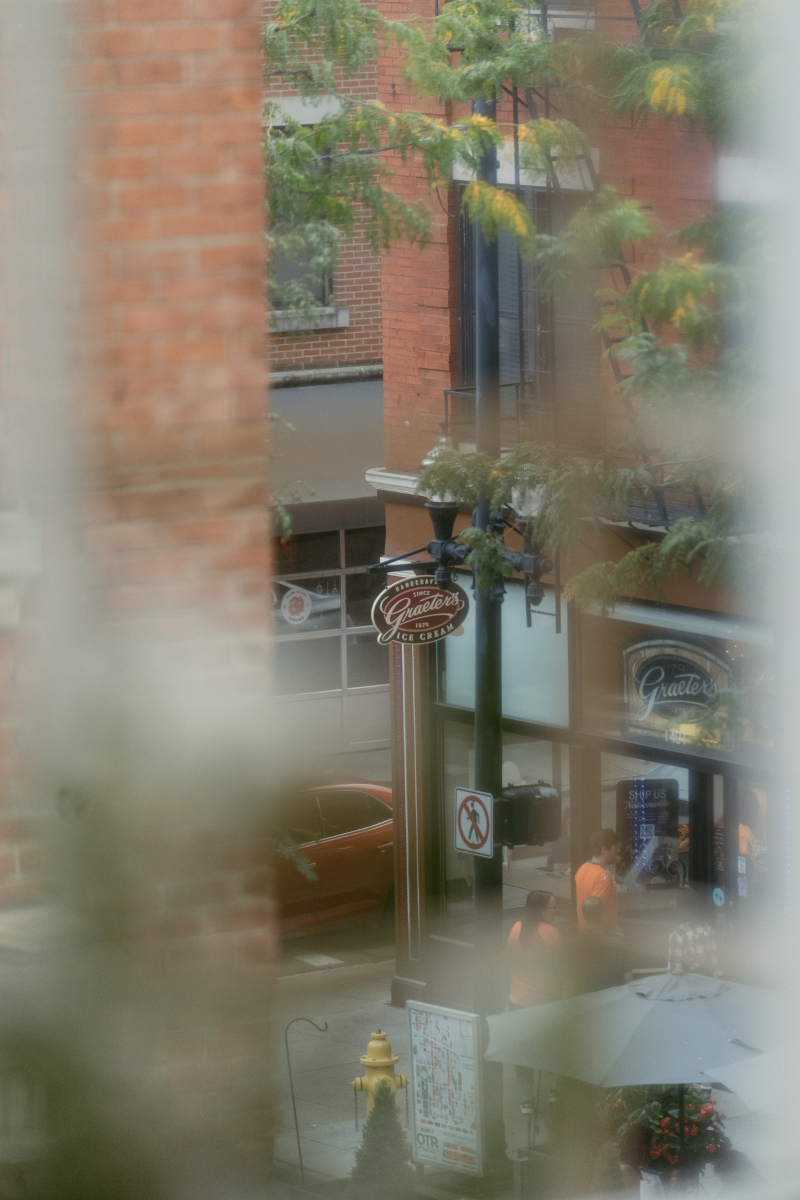 A view through a window showing a Graeter's Ice Cream shop on a city street, with people walking by and a red car parked outside. The photo is softly blurred around the edges, creating a dreamy feel.