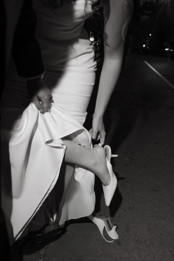 The bride slips off her heels on the sidewalk at night, holding up her dress with one hand.