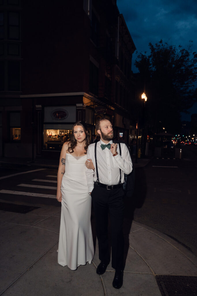 The bride and groom pose on a city sidewalk at night, looking off into the distance. She wears a sleek white gown, and he’s in suspenders with his jacket slung over his shoulder.