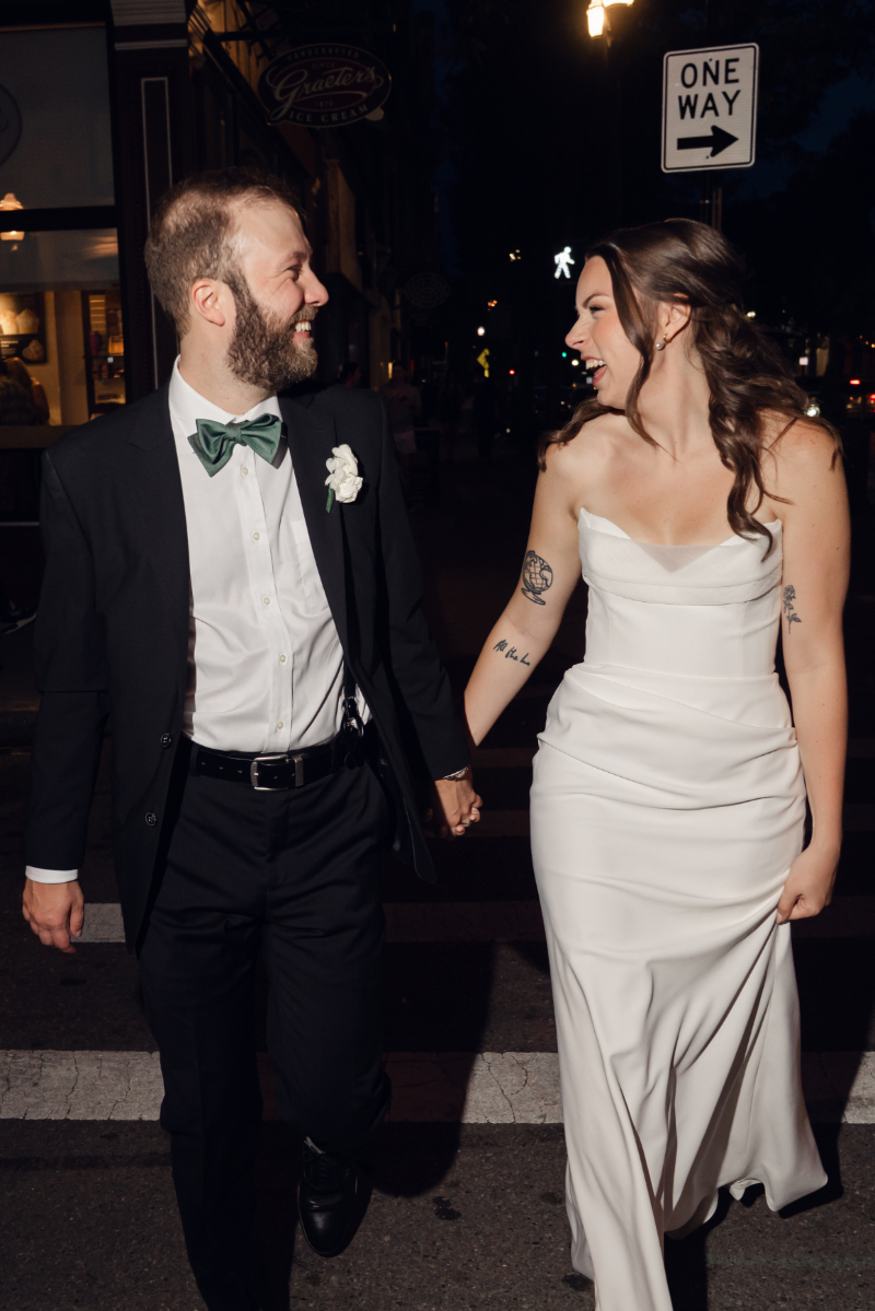 The bride and groom walk hand in hand at night, smiling and laughing as they look at each other. They both appear joyful and relaxed after their celebration.