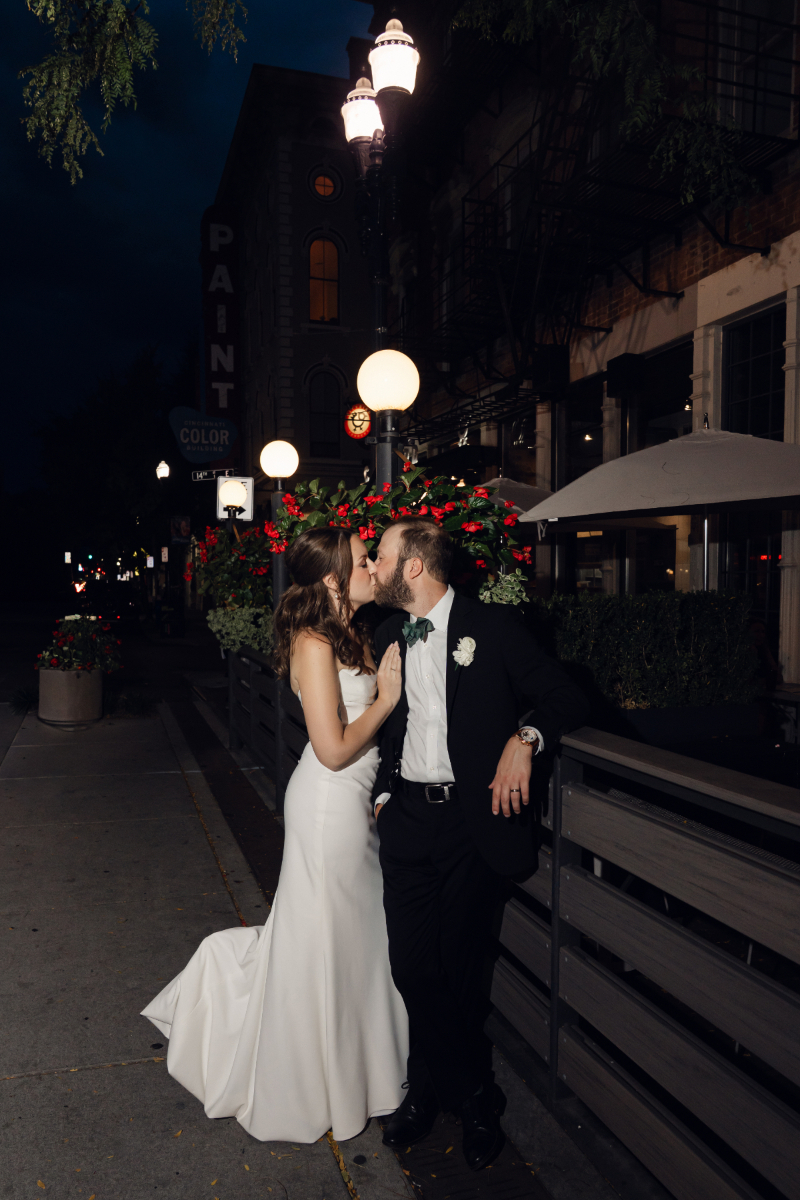 The bride and groom share a kiss on a quiet city sidewalk at night, surrounded by planters with red flowers and soft street lighting. The bride wears a sleek white gown, while the groom leans casually against a railing in his tuxedo.
