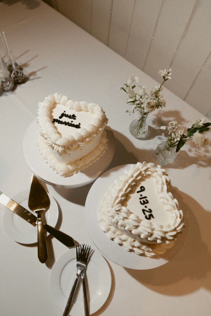 Two heart-shaped white cakes with piped frosting sit on a dessert table. One is labeled "just married" and the other features the wedding date "9.13.25." Small vases with white flowers and dessert utensils are also on the table.