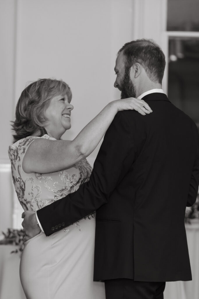 A black and white photo of a groom dancing with his mother during the wedding reception. They are smiling warmly at each other, sharing a joyful moment.