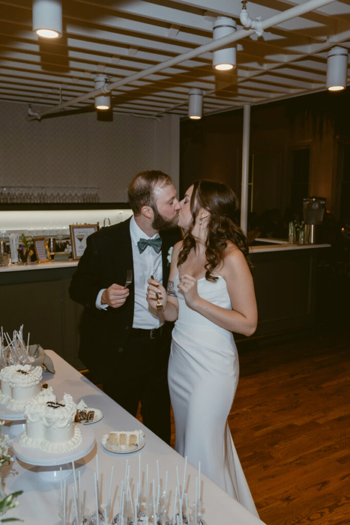 The bride and groom share a kiss during their cake cutting at the reception. They each hold a fork with a bite of cake, standing beside a dessert table with small cakes and treats.