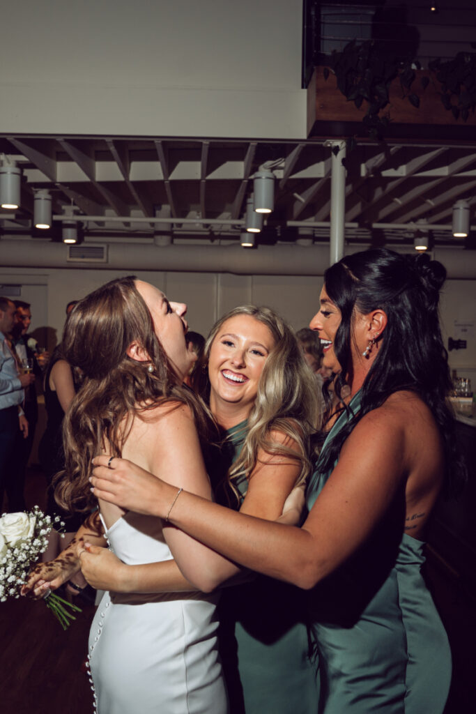 The bride laughs and embraces two bridesmaids on the dance floor. All three women are smiling and holding each other in a joyful group hug, clearly enjoying the celebration. The bridesmaids are wearing sage green dresses.