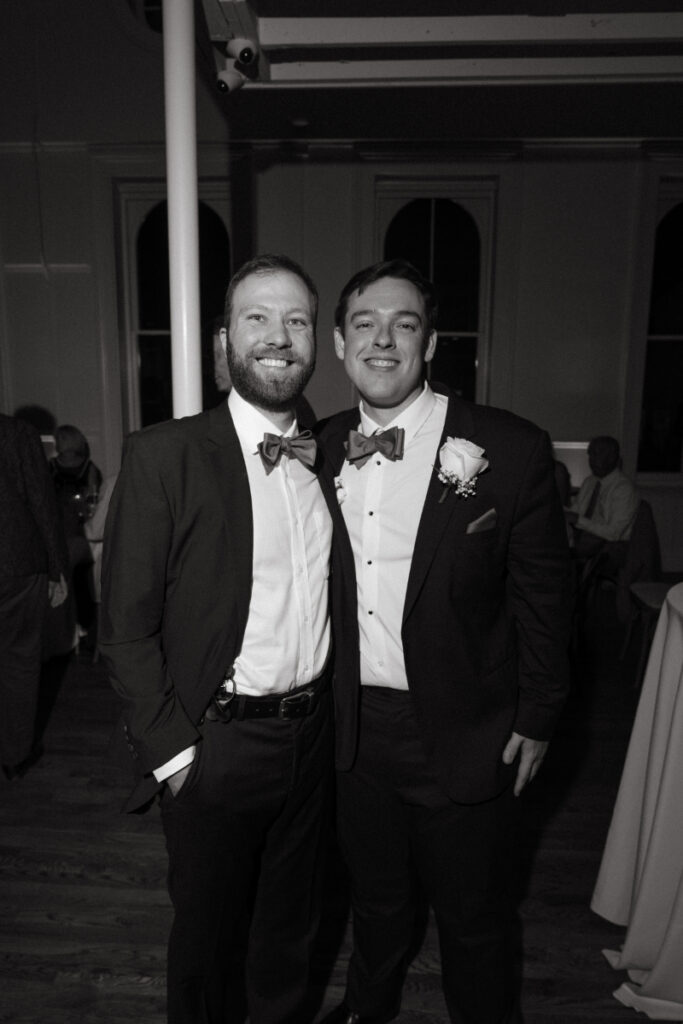 The groom smiles and poses with a groomsman at the reception. Both are wearing black suits with bow ties and boutonnières, standing close together in a warmly lit venue.