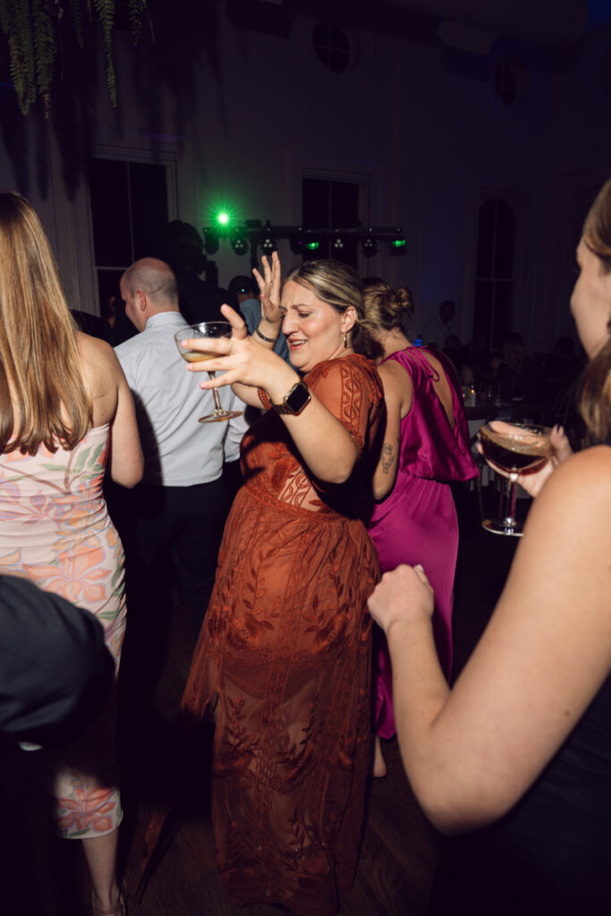 A guest in a burnt orange lace dress dances joyfully at the reception, holding a drink in one hand with her other arm raised. Other guests around her are also dancing, and the room is dimly lit with colorful party lights in the background.