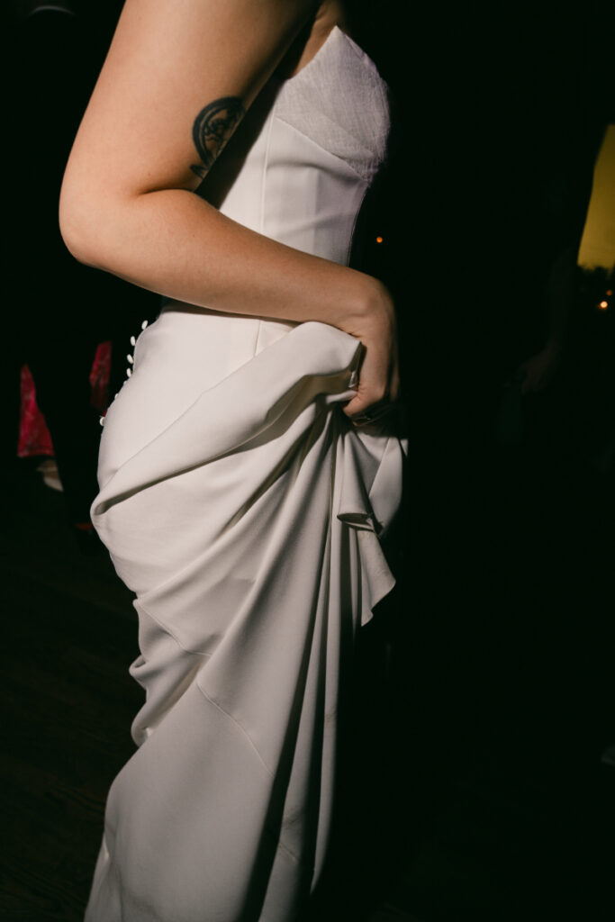 A bride holds up her sleek white gown slightly as she moves on the dance floor, the lighting highlighting the fabric and a tattoo on her upper arm.