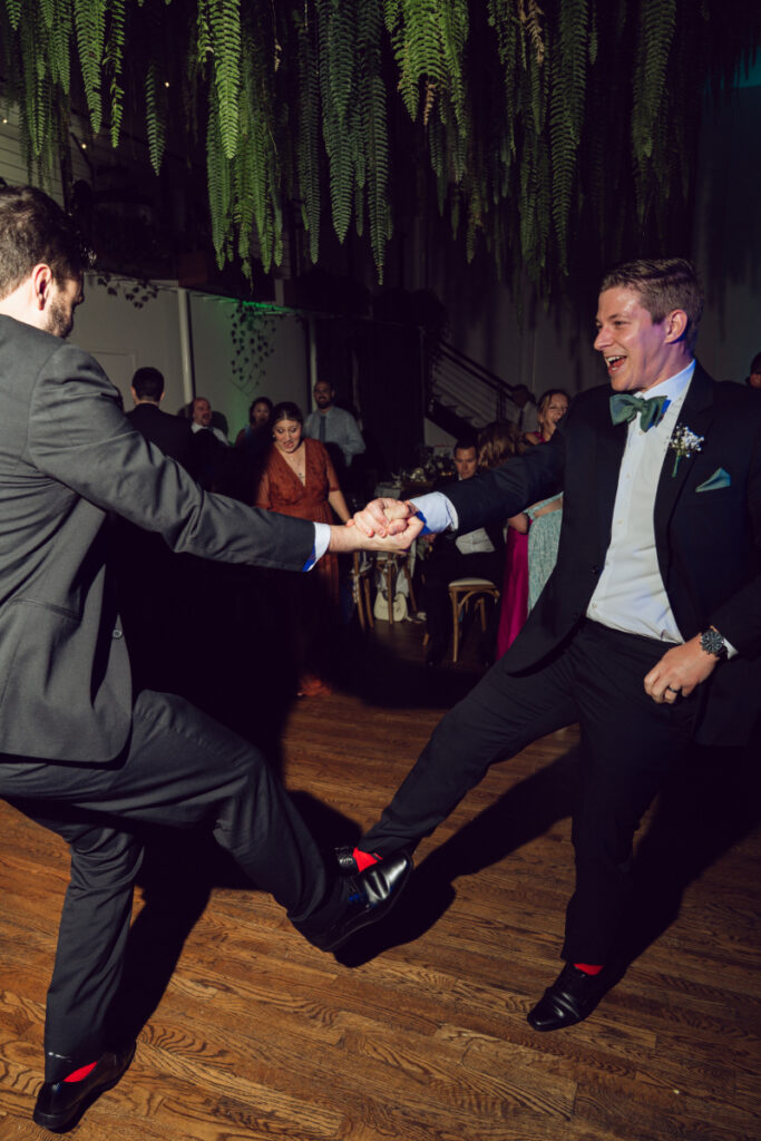 Two guests in tuxedos dance enthusiastically on the reception floor, holding hands and kicking their legs out in sync. Both are smiling and wearing fun red socks. The space is warmly lit with hanging greenery overhead and other guests seated in the background.