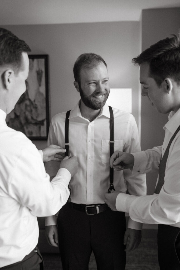 Groom smiling as groomsmen help adjust his suspenders before a Downtown Cincinnati Wedding.