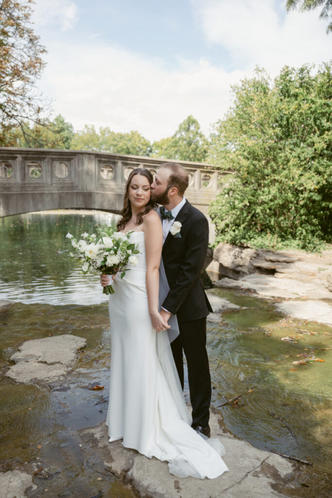 Groom kisses bride’s temple as they stand by the water during their Downtown Cincinnati Wedding.