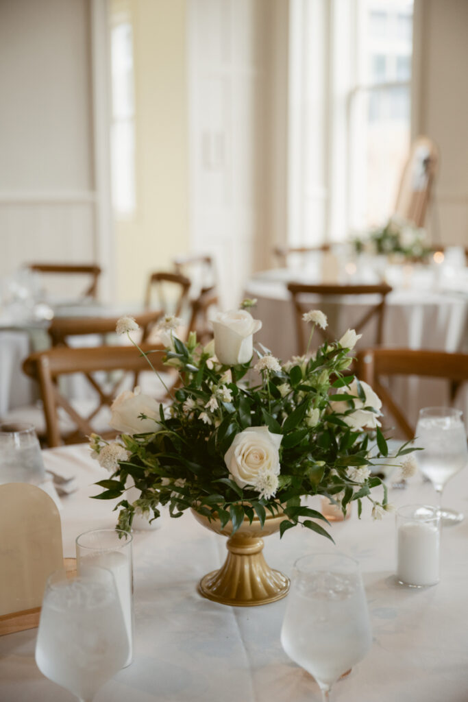 Gold compote centerpiece with white roses and greenery on a round reception table with candles.