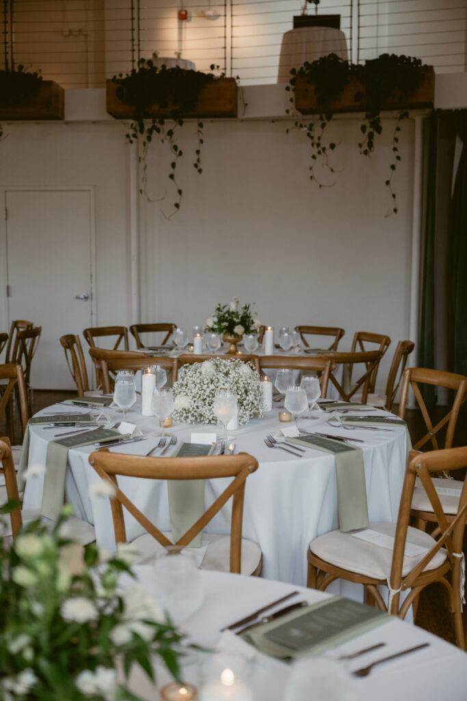 Reception tables with white linens, sage green napkins, and floral centerpieces, surrounded by wooden cross-back chairs in a softly lit room with hanging greenery.